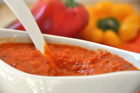 Crema di peperoni bimby in una ciotola bianca, con peperoni rossi e gialli sfocati sullo sfondo.