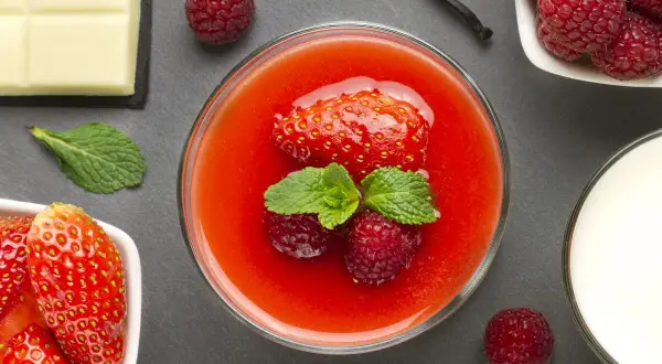 Un bicchiere di coulis di fragole con fragola, lamponi e menta. Accanto, fragole fresche, cioccolato bianco, latte e un baccello di vaniglia.