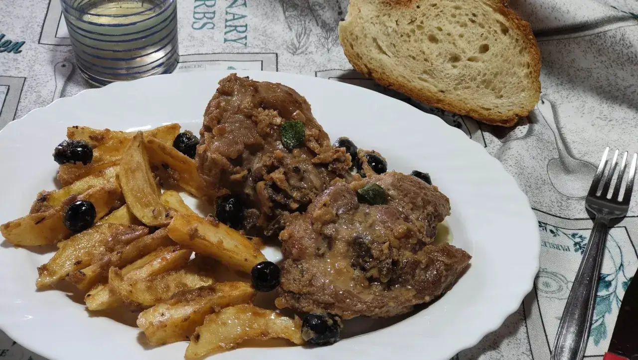 Un succulento ossobuco di tacchino con patate arrosto e olive nere, accompagnato da pane fresco e un bicchiere di vino bianco.