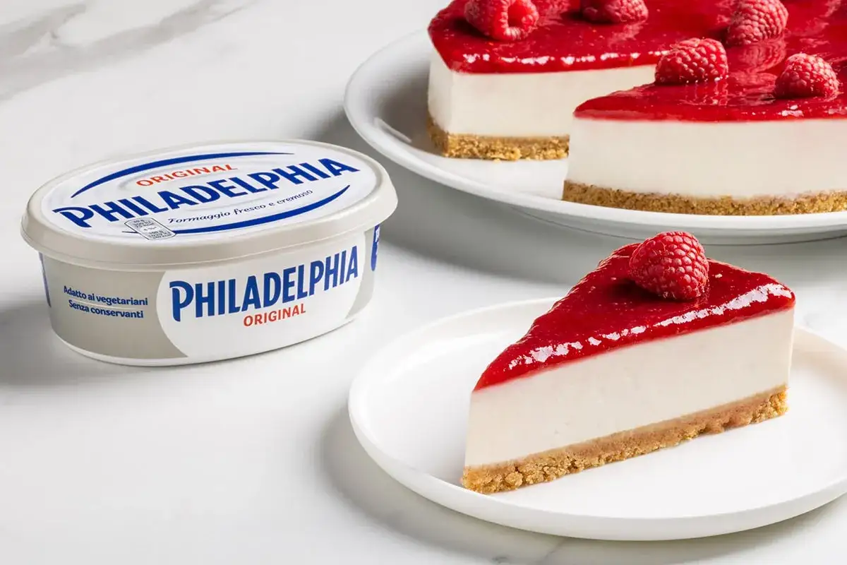 Cheesecake ricetta semplice con Philadelphia: una fetta cremosa con topping ai lamponi e un barattolo di formaggio fresco.