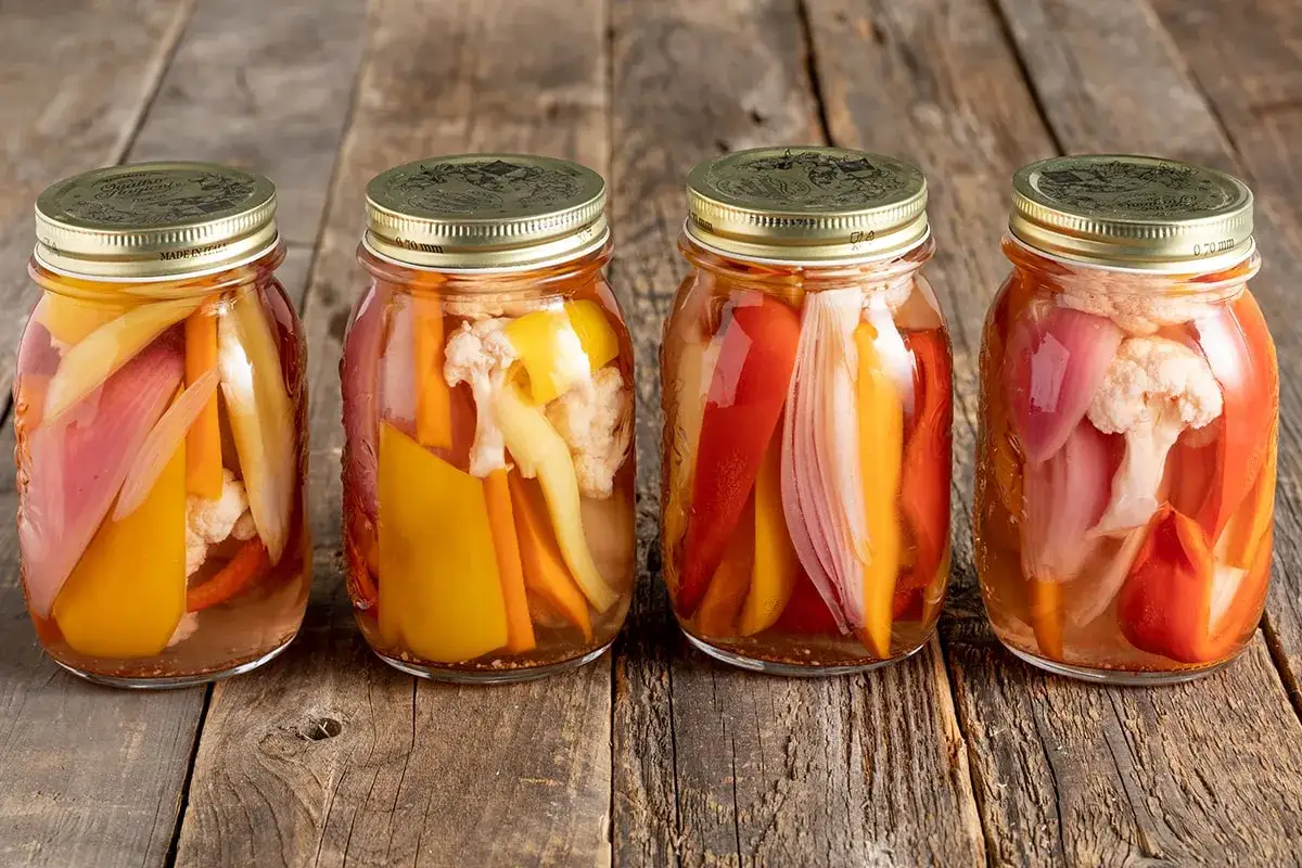 Quattro barattoli di giardiniera fatta in casa, con verdure colorate come peperoni, carote e cavolfiore, su un tavolo di legno.