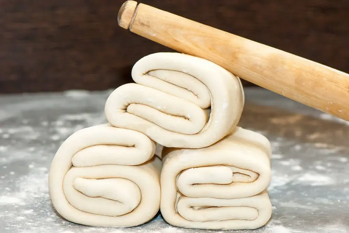 Tre blocchi di pasta sfoglia arrotolata, pronti per una ricetta pasta sfoglia veloce. Un mattarello in legno riposa sopra.