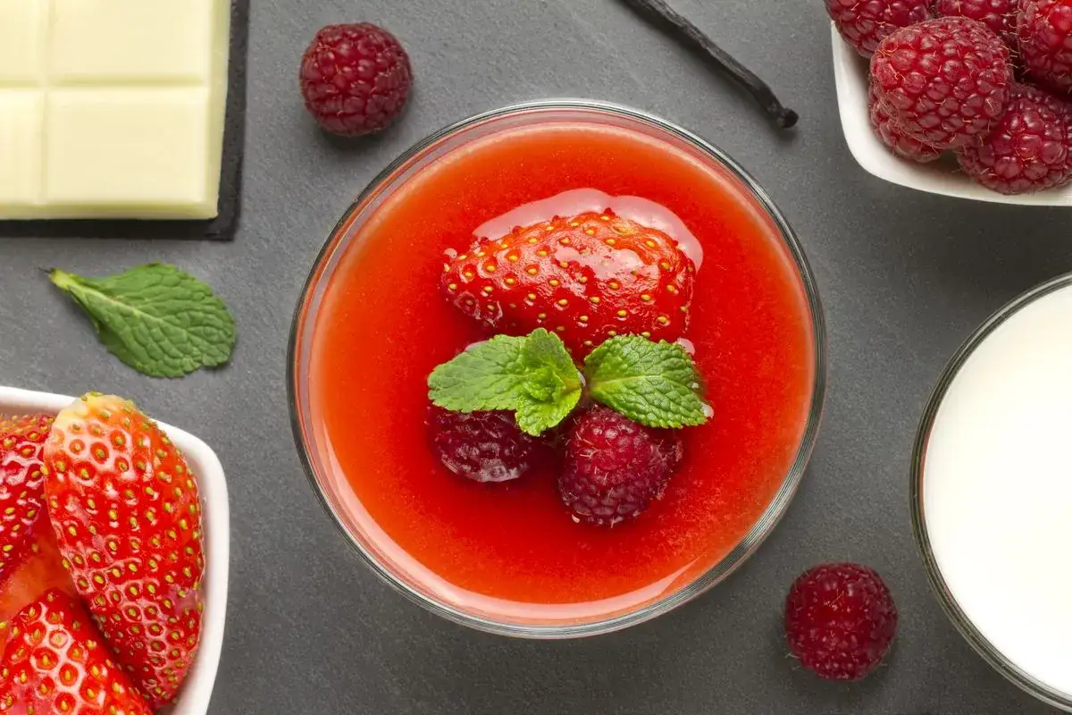 Un bicchiere di coulis di fragole, guarnito con fragola, lamponi e menta. Accanto, lamponi, cioccolato bianco, bacca di vaniglia e un bicchiere di latte.