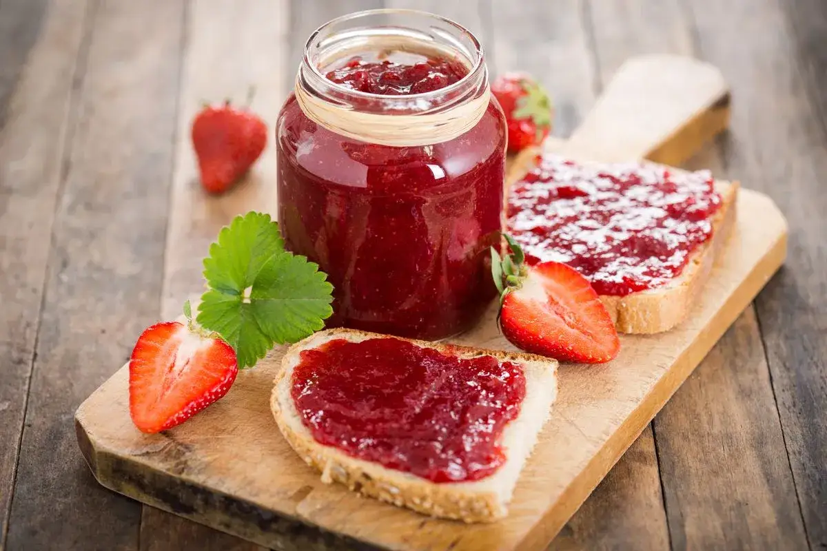 Marmellata di fragole fatta in casa con il Bimby, spalmata su fette di pane tostato, con fragole fresche e foglie verdi.